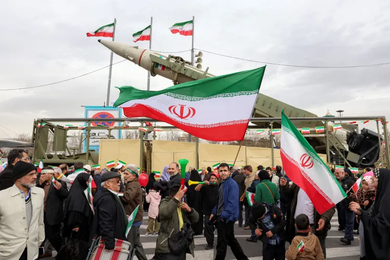People walk with Iranian national flags near a ballistic missile launch vehicle in Tehran on February 11, 2026, during a rally marking the 47th anniversary of the 1979 Islamic revolution. The Persian calendar date of Bahman 22 celebrates the anniversary of the resignation of the ousted shah's last prime minister and the formal assumption of power by revolutionary leader Ayatollah Ruhollah Khomeini. (Photo by AFP)